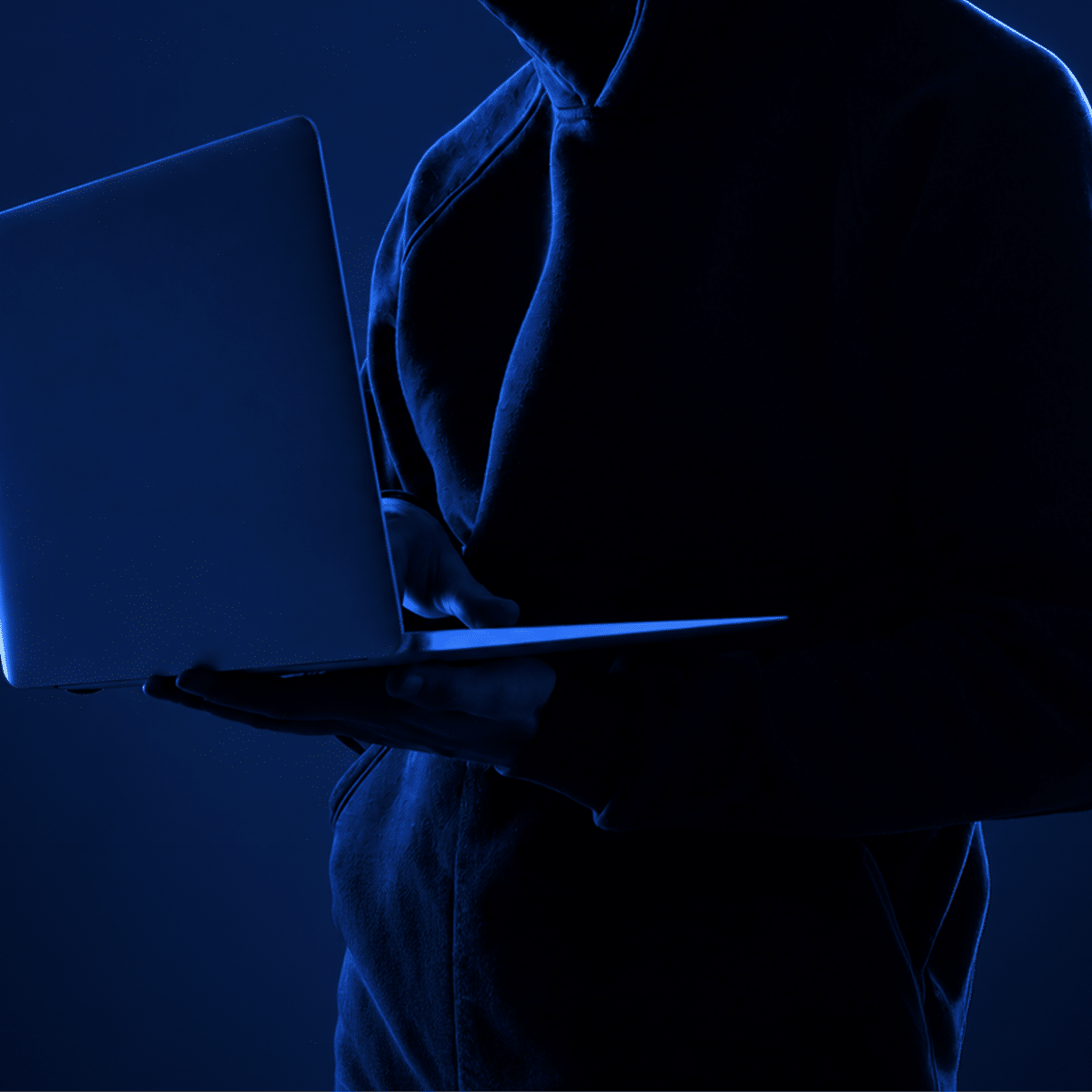Hooded Hacker stands in dark room holding a laptop highlighted by deep LED lighting.